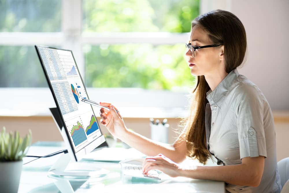  A person sitting at a desk, pointing to a computer screen displaying case management statistics. The screen shows various charts and tables, including bar graphs, pie charts, and line graphs, representing different data and trends related to case management. The person uses a pen to highlight specific areas on the screen, indicating analysis or review of the statistics.