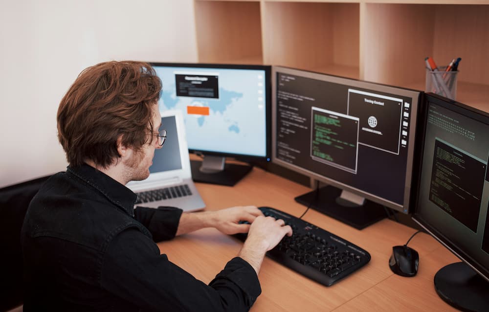 A professional workspace featuring multiple computer monitors displaying interfaces of case management software, including a world map, coding windows, and text boxes. A person is typing on a keyboard, with a laptop and stationery neatly arranged on the desk.