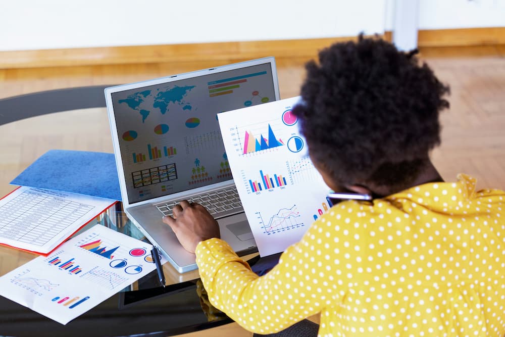  A person dressed in a yellow polka dot shirt sits at a desk working with data management software displayed on their laptop screen. The software showcases a variety of visualizations, such as world maps, bar graphs, pie charts, and line graphs. The person holds a sheet with similar data visualizations while additional sheets featuring charts and graphs are spread out across the table.