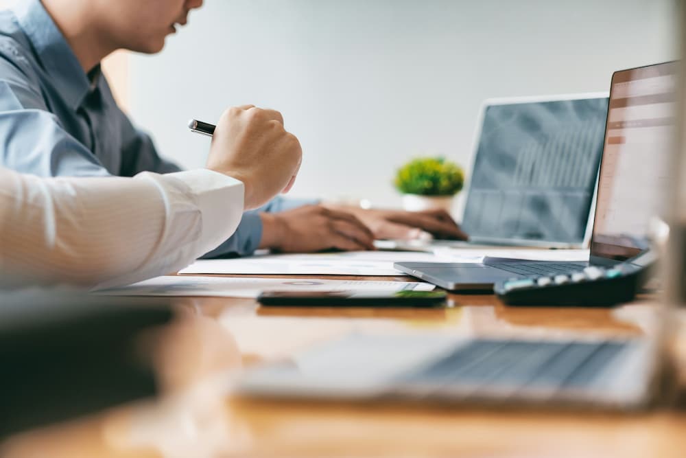 Two individuals working at a desk covered with laptops, papers, and a calculator, reflecting a professional setup where data management software is being used to organize and analyze information effectively.