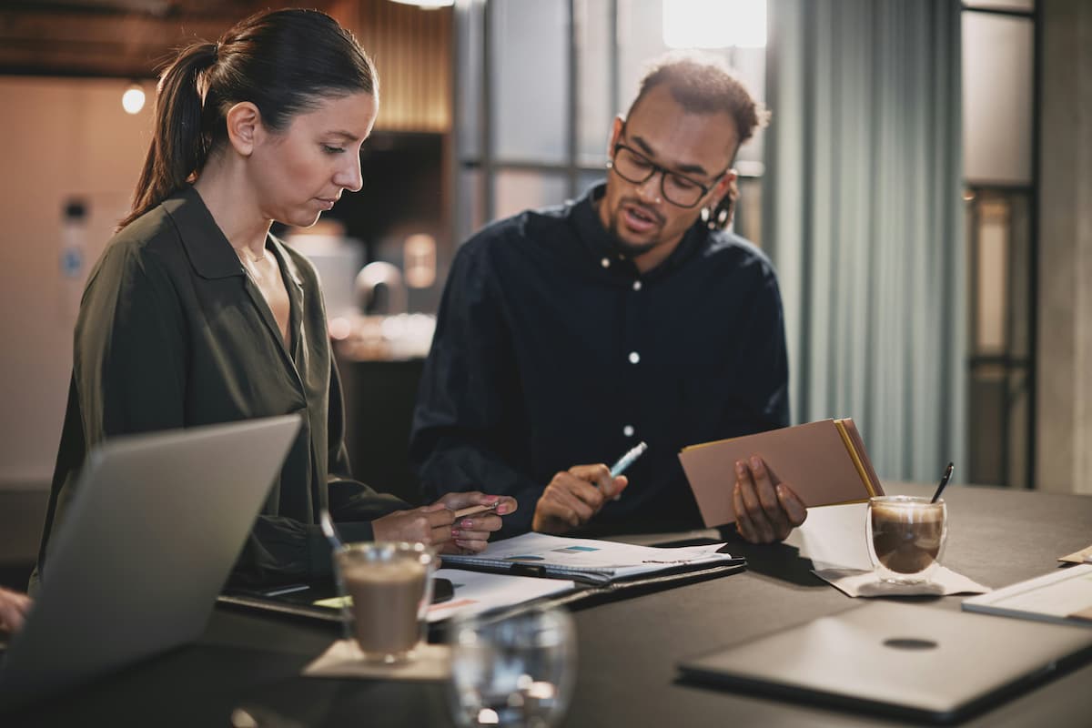 Two professionals discussing data at a table with laptops and charts, analyzing strategies to boost custom software ROI.