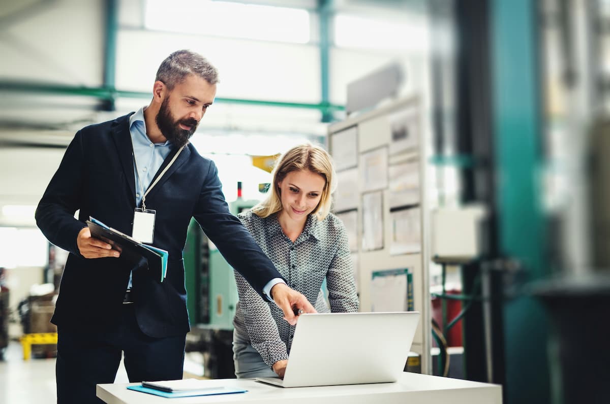 Managers reviewing a manufacturing analytics dashboard on a laptop while discussing performance in a factory environment.