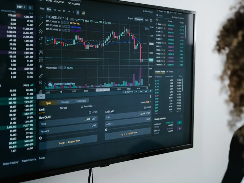 This image shows a person analyzing a cryptocurrency trading platform displayed on a large monitor. The screen is filled with various types of financial data, including candlestick charts, order books, volume indicators, and trading options.