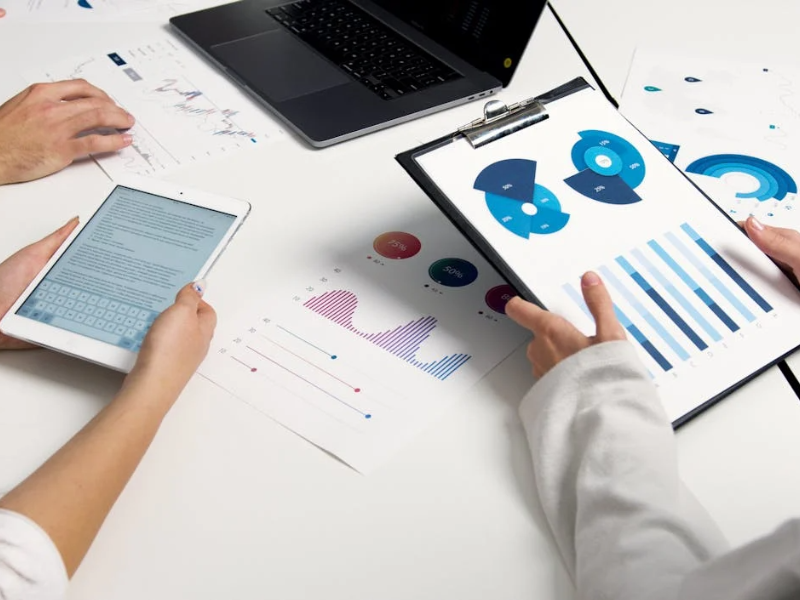 This image shows a group of people working at a desk analyzing various charts and data. One person is holding a clipboard with colorful pie charts and bar graphs, while another is using a tablet that displays a block of text, possibly a report or analysis.