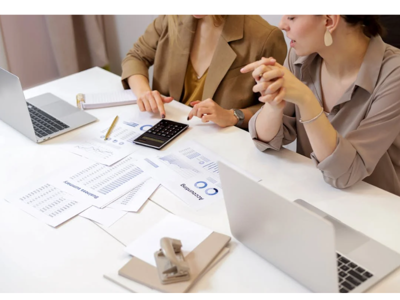 The image shows two professional women working at a modern office desk, engaged in a financial or accounting discussion.