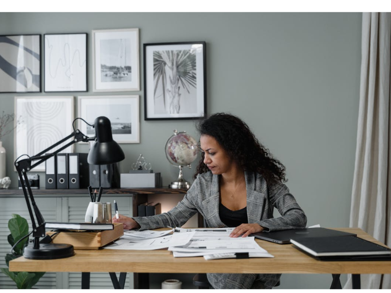 The image shows a professional woman working at a stylish office desk, deeply focused on reviewing documents and paperwork.