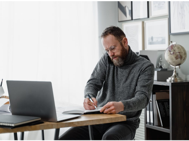 The image shows a man working in a modern office space, multitasking as he speaks on the phone and takes notes in a notebook.