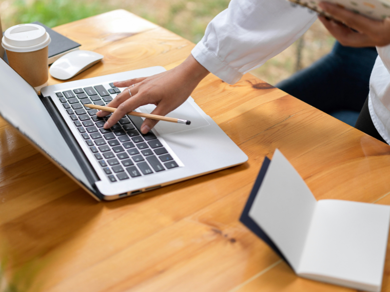 The image shows a person working at a wooden desk with a laptop computer. The setting appears to be a casual or semi-professional workspace, possibly outdoors or near a window with natural light.
